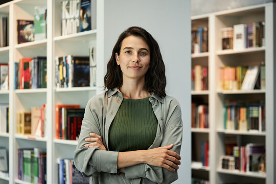 Horizontal medium portrait of confident young Caucasian woman wearing casual clothes standing with arms crossed against bookshelves in library looking at camera
