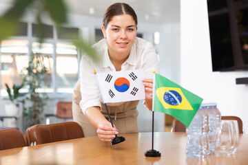 Positive young woman putting little flag of South Korea on the table with flag of Brazil in...
