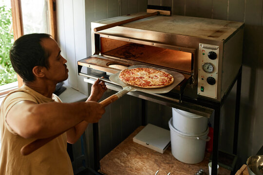 Horizontal medium shot of mature man working in kitchen taking out pizza from oven using bakers shovel