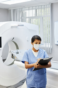 Vertical high angle shot of unrecognizable radiographer wearing mask on face working in modern hospital standing against MRI scanner holding clipboard making notes