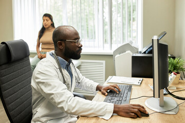 Horizontal medium portrait of African American man working on computer in modern hospital, his patient sitting on background