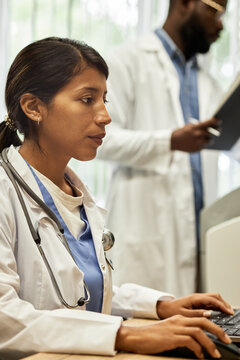 Vertical medium portrait of serious Hispanic woman working on computer in modern hospital, her African American colleague doing paperwork on background