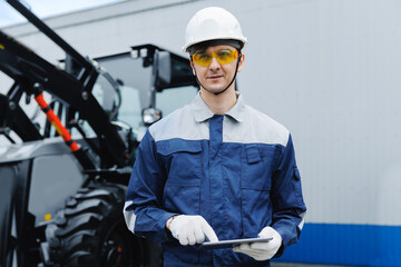 Male worker in mining industry wearing safety gear holding tablet on background tractor