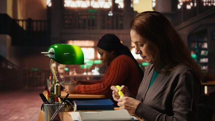 Mature woman student in the library highlighting important ideas for exams, focusing on her studies surrounded by academic materials and books. Study strategy memorization.