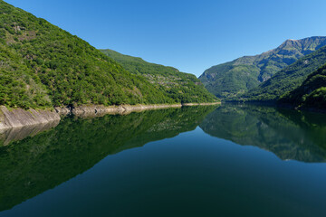 serene view of a tranquil Swiss lake surrounded by lush green mountains under a clear blue sky,...