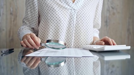 Auditor woman analyzing financial documents with magnifying glass and calculator at her office desk...