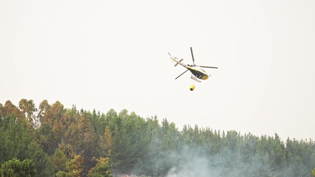 Aerial firefighting helicopter hovering above burning forest, strategically dropping massive water load from specialized bambi bucket to combat spreading wildfire