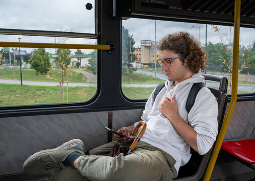 Prague, Czech Republic, August 6, 2023. A young Caucasian man with curly hair and glasses is sitting at the bus terminal. He's waiting for the bus to depart, using his cell phone. It's a rainy day.