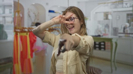 Woman points sunglasses to camera in a bright clothing store with racks; playful joy excitement spontaneity.