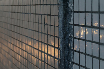 Close-up of tiled urban wall showing worn texture, mesh shadow diagonals, and golden sunlight...
