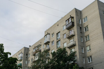Low-angle view of gray brick residential building with varied balconies, white-framed windows, green trees, and overhead power lines under cloudy sky.