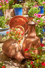 Two antique copper jugs on a sunny patio surrounded by colorful flowers in clay and ceramic pots,...