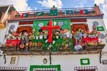 A Spanish building balcony decorated with a large red floral cross, colorful flower pots, guitars,...