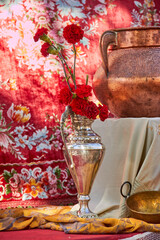 A silver vase with red carnations stands beside a large copper pot against a floral patterned backdrop, creating a rich and traditional composition. still life