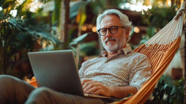 Mature man with gray hair and glasses relaxing in a hammock outdoors while working on a laptop surrounded by lush greenery and warm natural light