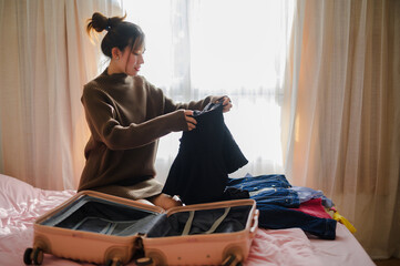 A young woman is packing clothes into a suitcase, sitting on a bed in a well-lit room, with sunlight streaming through the window.