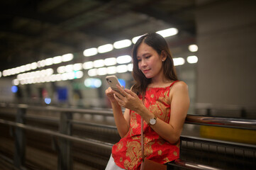 A young woman in a vibrant red top is focused on her smartphone, standing in a well-lit transit...