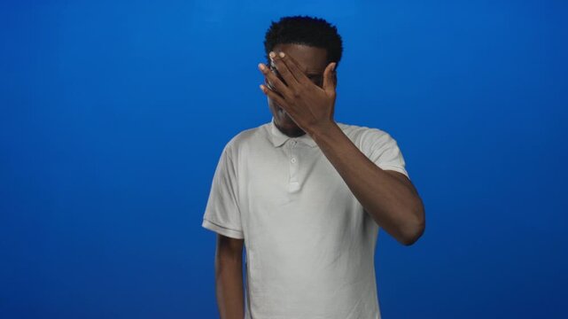 Young african american man with outstretched palm blocking the camera view in a bright blue studio environment; defiance determination.