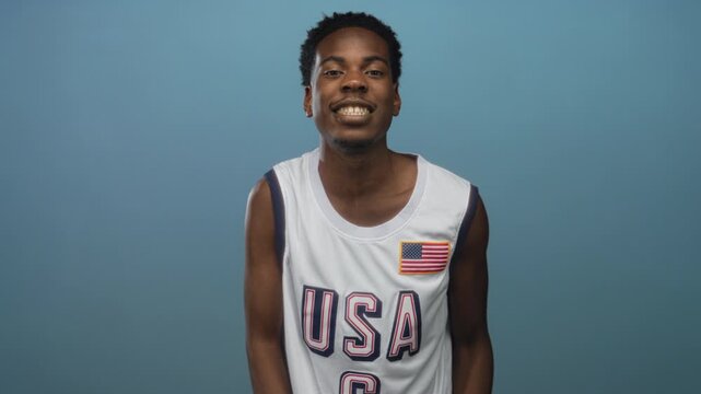 Man wearing usa basketball jersey with american flag patch smiles and raises hand in pale blue studio; confidence.