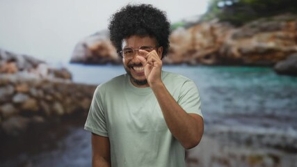 Smiling man with curly hair in glasses making small gesture at seaside with rocks and ocean in background.