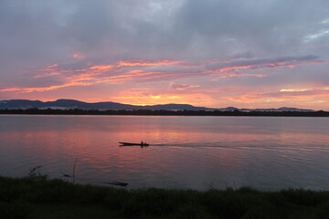 Fototapeta premium A breathtaking sunset over a calm river with vibrant orange and purple hues painting the sky. A small boat glides across the water, leaving gentle ripples under the silhouette of distant mountains.