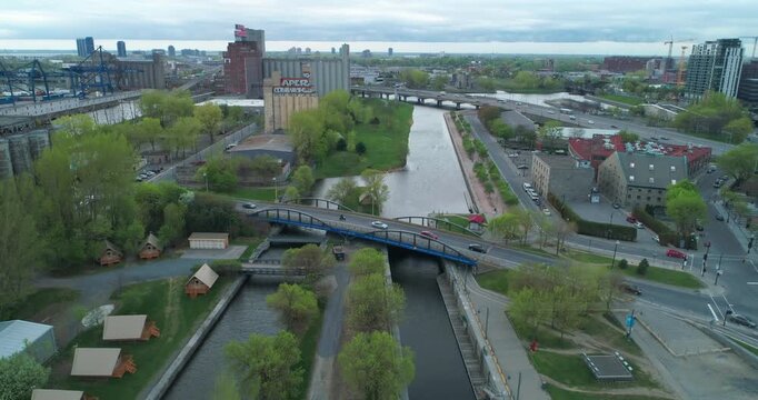 Aerial view of Lachine Canal in Montreal, Canada. Cars and a motorcycle cross a blue bridge over the canal. Industrial buildings and city skyline in background.