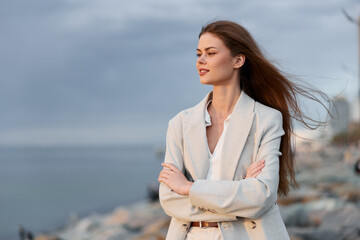 confident woman in beige blazer standing near sea coast with crossed arms natural light autumn...