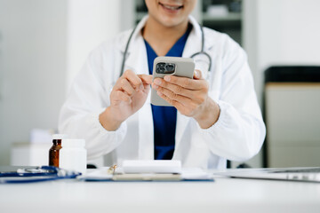 Male doctor in uniform using smartphone at medical office desk with pills, clipboard, and...