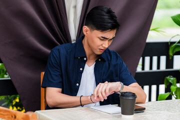 Asian man in casual navy shirt frowns at his wristwatch at a garden café table with coffee,...