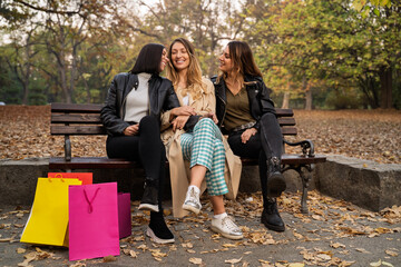 Three beautiful girls in twenties sitting on park bench with shopping bags and smiling, autumn...