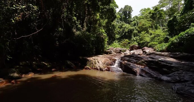 Cascading water flows over smooth boulders at Lubuk Teja waterfall, hidden in lush Tioman Island rainforest. Calm brown pond in shadow contrasts with bright, sunlit stream and greenery above.