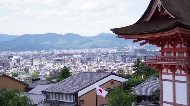 Panoramic view of Kyoto city with the Japanese flag in the foreground and a traditional temple roof to the right. Lush green mountains frame the skyline, blending modern urban scenery with Japan&rsquo;s ric