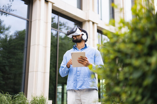 An Indian man wearing a VR headset and headphones holds a tablet outside a modern building, possibly for augmented reality. - Powered by Adobe