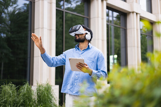 A man wearing a VR headset and holding a tablet gestures outdoors in front of a modern building with a green landscape.