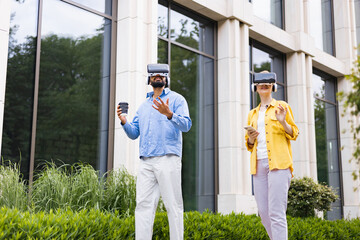 Two people wearing VR headsets explore a virtual reality experience outdoors in front of a modern building.