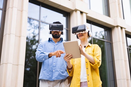 A diverse couple explores augmented reality using VR headsets and a tablet outdoors near a building. - Powered by Adobe
