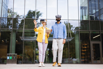 Two people wearing VR headsets walk in front of a modern glass building, symbolizing technology and innovation in an urban environment.