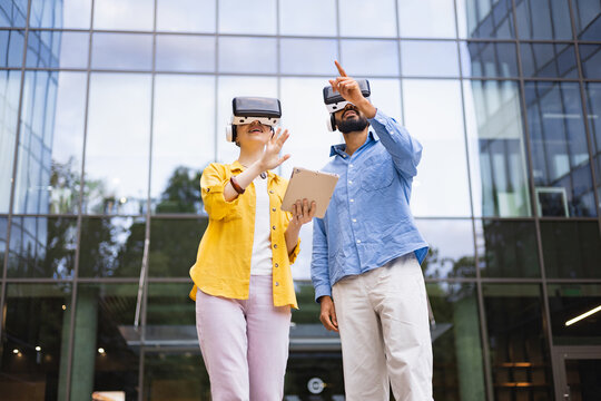 Two people wearing VR headsets in front of a modern building explore a virtual environment together, one holding a tablet.