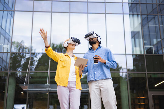 A diverse couple wearing VR headsets explore a futuristic experience outdoors, with a tablet and coffee in hand, in front of a modern building.