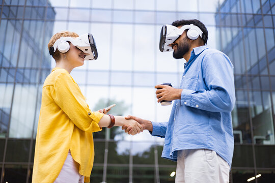 Two people wearing VR headsets shaking hands in front of a modern building, a coffee cup present.