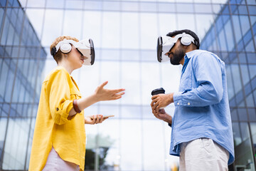 Two people wearing VR headsets interact outside a modern glass building, exploring virtual reality with coffee and a phone.