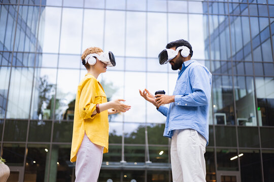 A diverse pair, immersed in virtual reality, stands before a modern glass building, engaged in what appears to be a digital interaction.