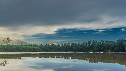 Early morning in the jungle. Thickets of rain forest on the riverbank. Clouds in the blue sky. Reflection on the smooth shiny surface of the water. Malaysia. Borneo. Kinabatangan River.