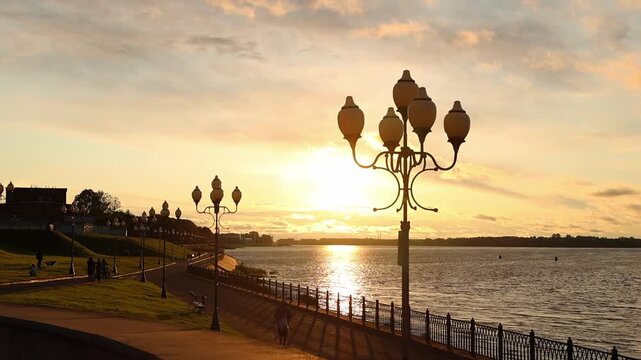 Picturesque Rybinsk riverside promenade at dusk, featuring decorative lamp posts, tranquil waters, and a warm summer sunset. Golden light above Volga river in beautiful old town.