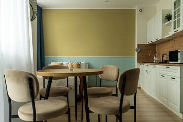 interior of a small apartment with bed, pillows, light kitchen furniture, chairs with a table, kitchen appliances and refrigerator. apartment has blue and brown. fruits on the kitchen table.