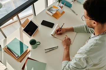 High angle view of college student sitting at his desk with digital tablet making notes in notebook and studying in the room