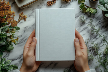 Woman hands holding white blank hardcover book on white marble table with natural plants and leaves. Top view to closed book, diary, planner mockup