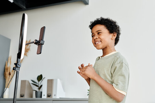 Horizontal image of young blogger talking on camera of his smart phone while standing in front of selfie lamp in the room