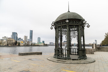 Wrought iron gazebo on the embankment of Iset River in Yekaterinburg, with modern city skyline and...