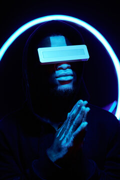 Vertical conceptual portrait of religious Black man with paint on face wearing hoodie and futuristic neon light eyewear standing in dark room with circle lamp praying to God